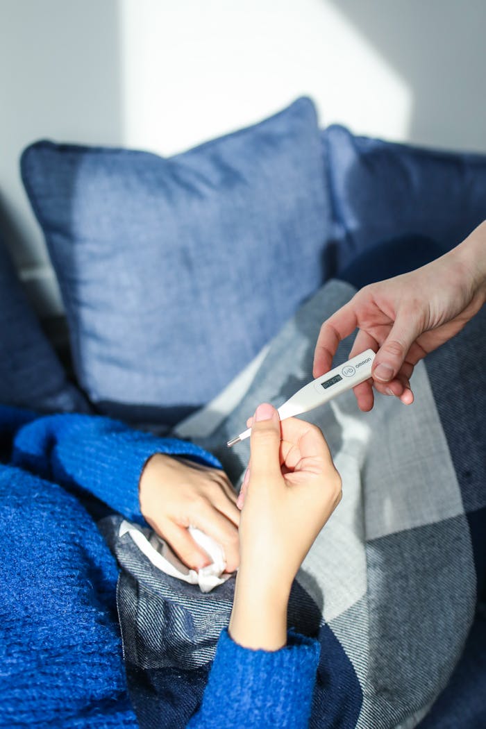 A woman checks her temperature using a digital thermometer while resting on a couch at home.